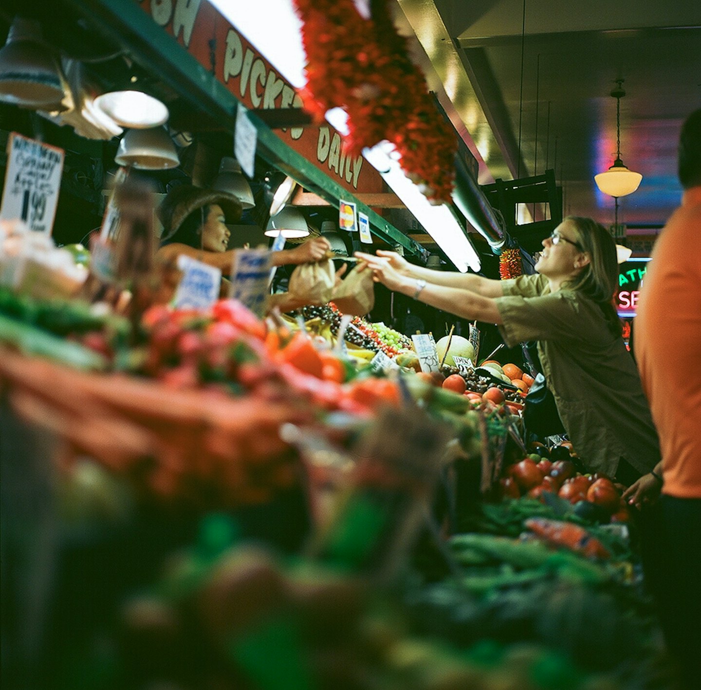 Vendor hands produce to a happy customer at a busy market stall.